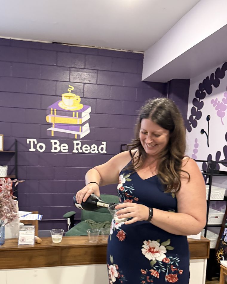 Jen Ostafinski, owner of To Be Read, smiling as she pours a glass of champagne at the shop counter, in front of the hand-painted To Be Read logo on the purple brick wall